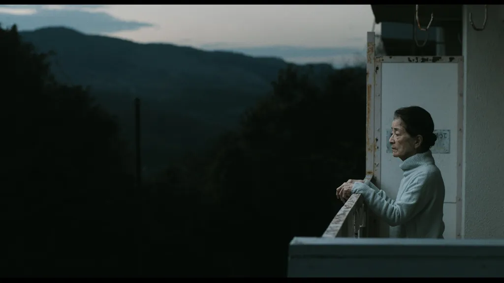 Chieko Baisho dans le film « Plan 75 », de Chie Hayakawa, 2022 (EUROZOOM). Une femme âgée s'appuie à la rambarde de son balcon, elle semble pensive et on voit la forêt et la montagne au loin.